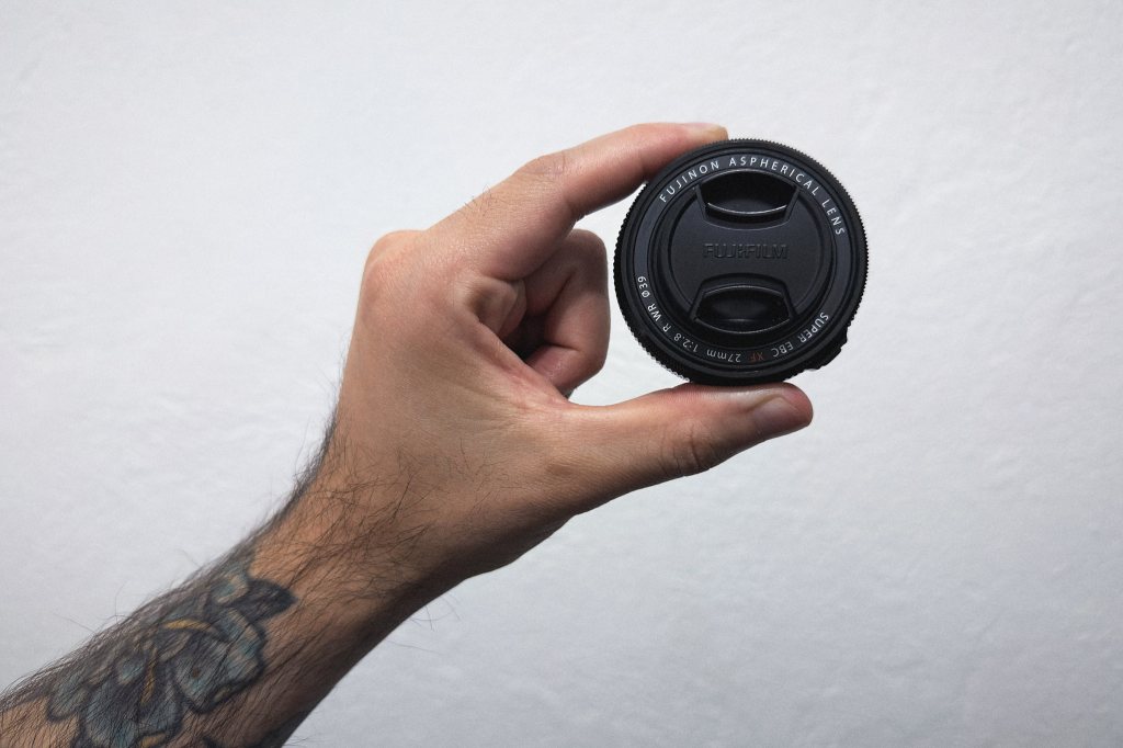 Mano sosteniendo el lente Fujinon XF27mm f/2.8 R WR sobre fondo blanco, mostrando su tamaño compacto