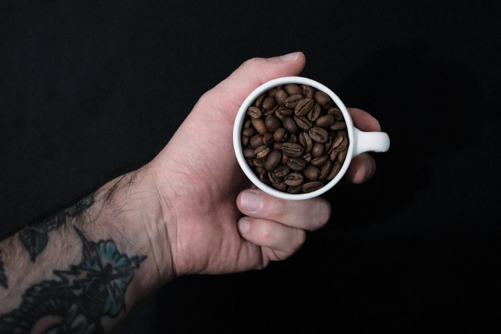 Mano sosteniendo una taza pequeña llena de granos de café sobre un fondo oscuro y desenfocado.