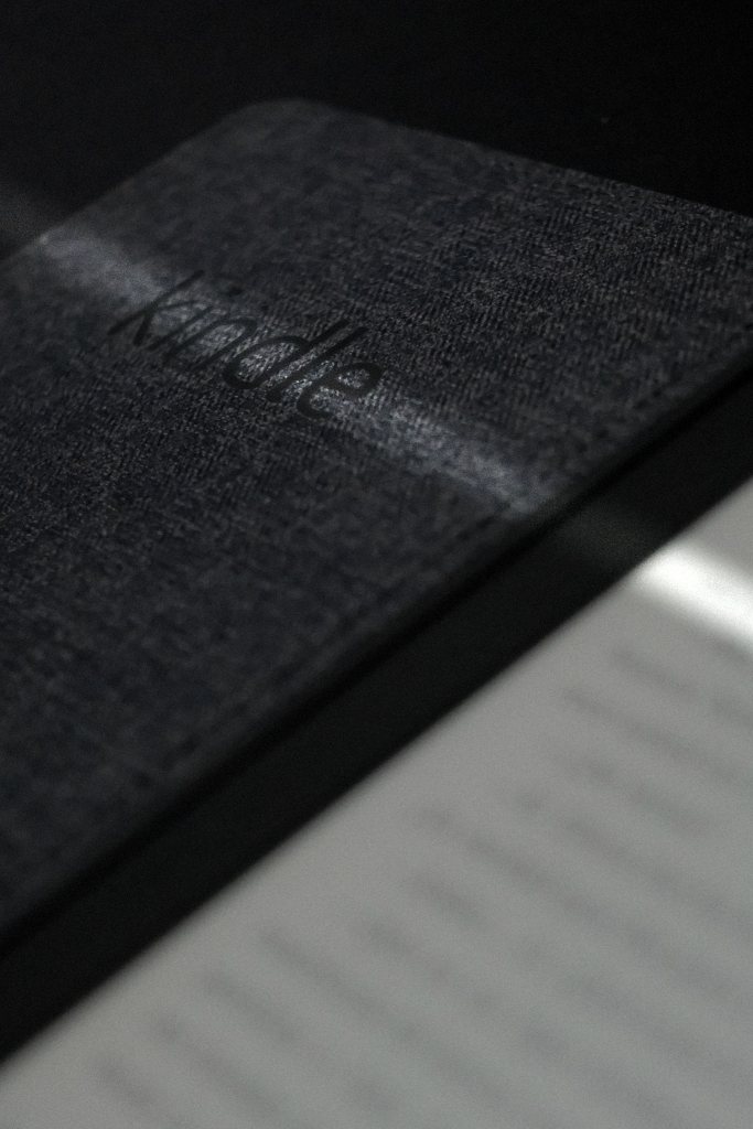 Detalle cercano de la funda texturizada de un Kindle con la palabra “kindle” en relieve, con una página desenfocada en primer plano y sombras suaves.