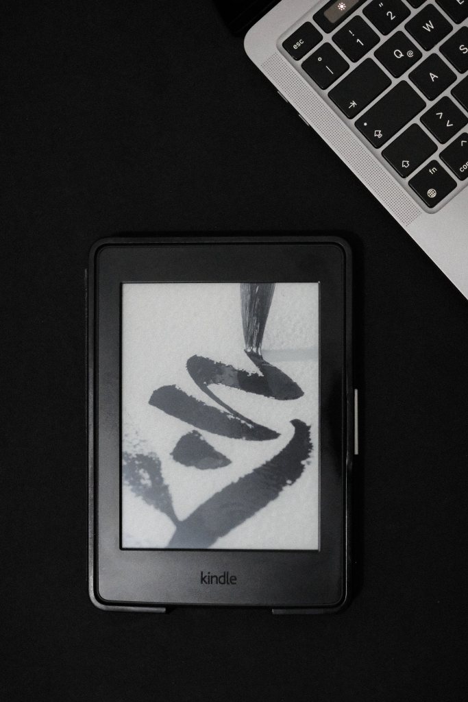 Vista cenital de un Kindle sobre una superficie oscura, con una laptop en la esquina superior derecha; en la pantalla del Kindle se ve una imagen en blanco y negro tipo caligrafía.