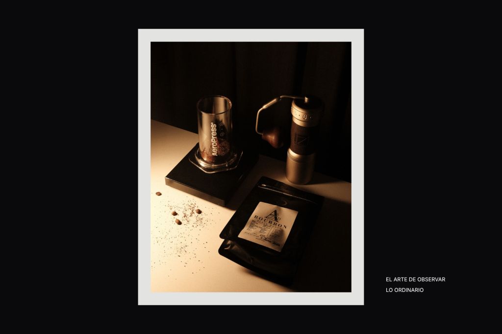 Fotografía editorial en formato horizontal con una imagen vertical al centro, enmarcada sobre fondo negro. La foto muestra una escena de café con una AeroPress, un molino manual, granos de café y una bolsa de café sobre una mesa iluminada con luz cálida lateral y sombras profundas. A la derecha aparece el texto: “El arte de observar lo ordinario”.