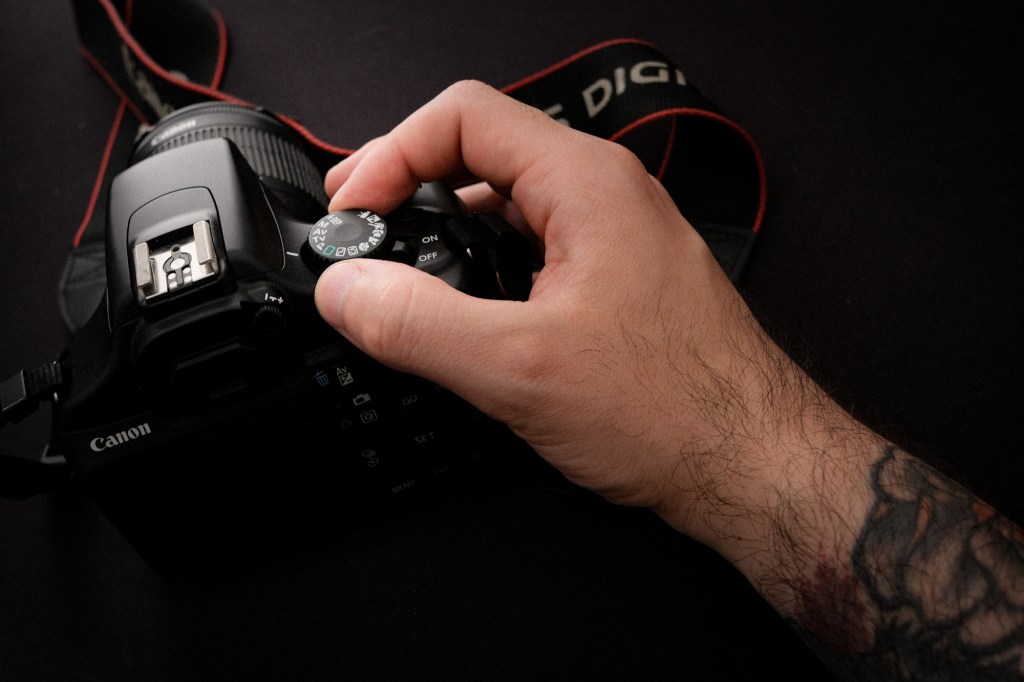 Un primer plano en ángulo de una mano tatuada (dedos índice y pulgar) ajustando el dial de modo de una cámara Canon DSLR negra (EOS Rebel T3). Se aprecian claramente los iconos de los modos de disparo en el dial. Fondo oscuro y texturizado.