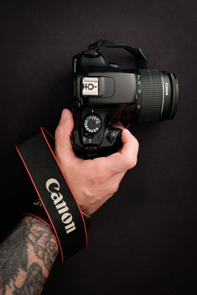 Una vista superior de una mano tatuada sosteniendo una cámara Canon DSLR negra (una EOS Rebel T3) con un objetivo 18-55mm adjunto. El dial de modo y el botón de disparo son visibles. Una correa negra de la marca Canon está enrollada alrededor de la muñeca. Todo sobre un fondo negro texturizado.