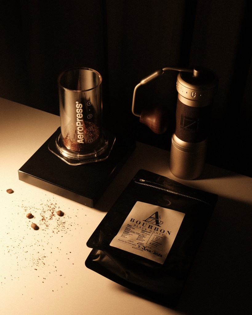 Escena de café con AeroPress, molino manual y una bolsa de café Bourbon Black Honey sobre una mesa iluminada con luz cálida y sombras marcadas.