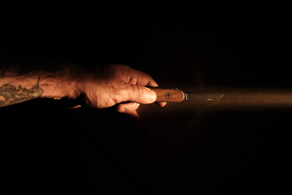 Plano detalle de una mano humana sosteniendo una navaja multiusos Victorinox con cachas de madera, iluminada por un haz de luz dramático sobre un fondo completamente negro. Imagen de portada para la newsletter Recortes número 001.