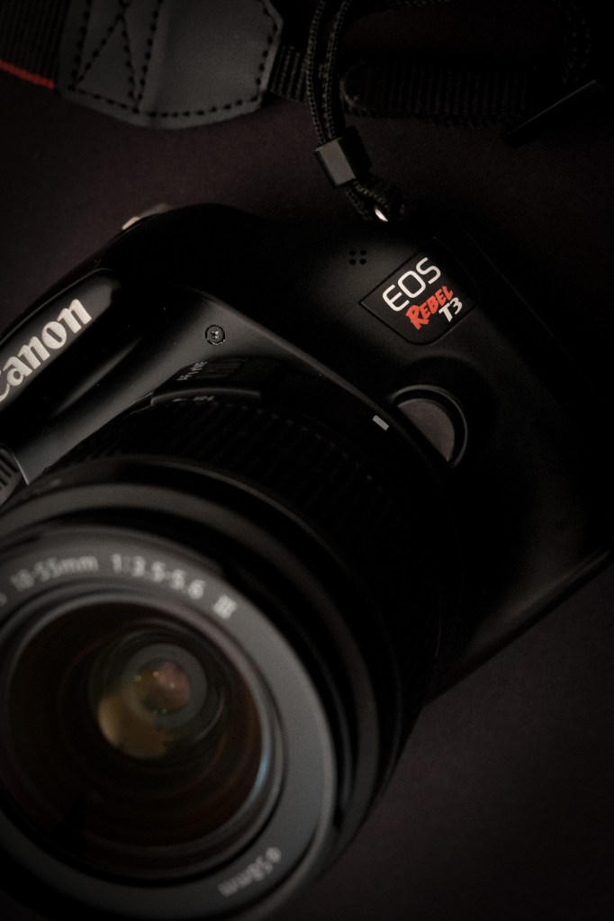 Un primer plano detallado e iluminado de manera dramática de una cámara Canon EOS Rebel T3 negra, enfocando nítidamente el distintivo "EOS Rebel T3" en blanco y rojo en el cuerpo de la cámara. El logotipo de "Canon" es visible cerca. Parte de la lente está en primer plano sobre un fondo oscuro.
