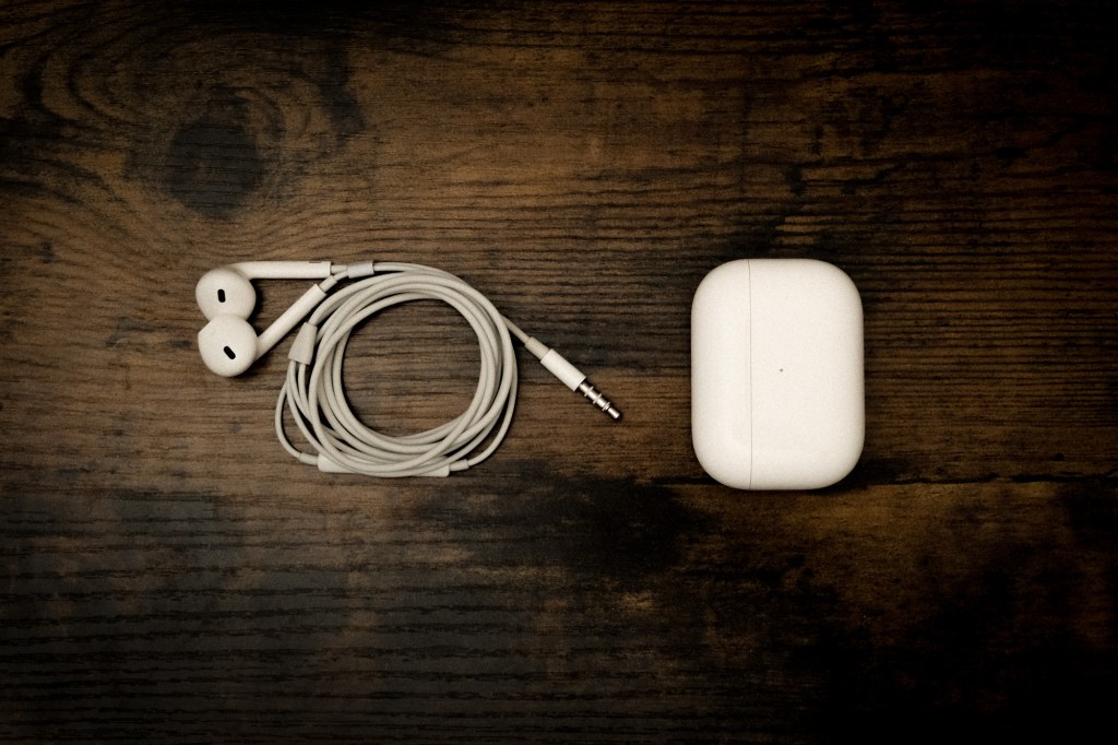 Fotografía cenital de unos audífonos blancos con cable junto a unos audífonos inalámbricos en su estuche, colocados uno al lado del otro sobre una mesa de madera oscura y avejentada con texturas marcadas.