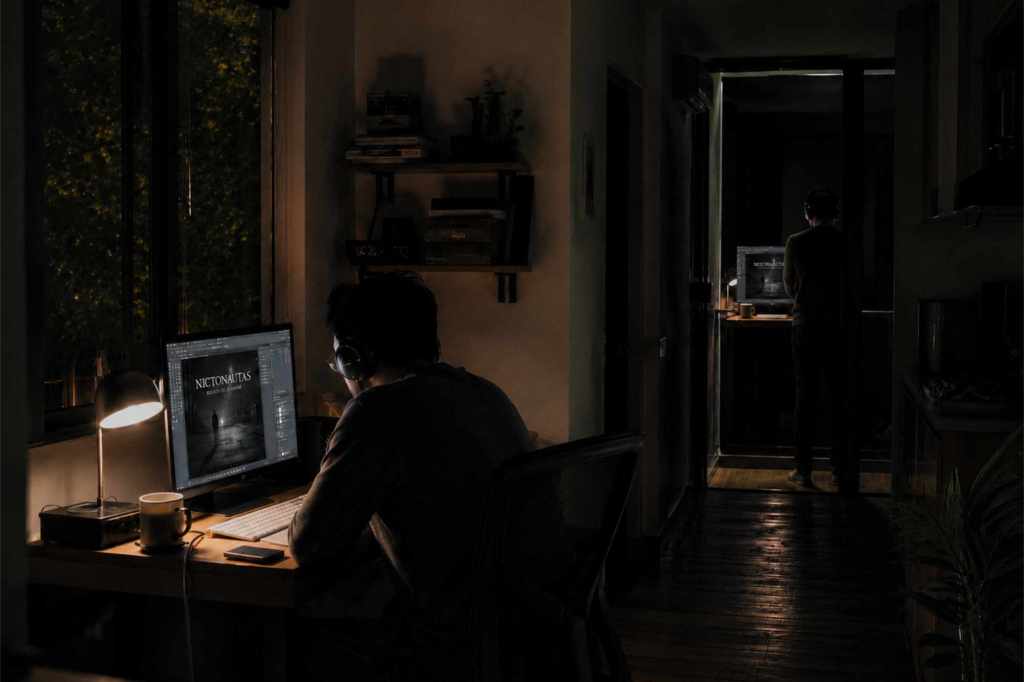 Hombre trabajando de noche frente a una computadora mientras su reflejo permanece de pie al fondo, frente a una puerta de cristal.